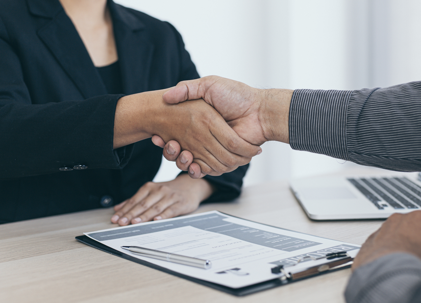What appears to be a woman and a man shaking hands over a desk with a resume and laptop visible