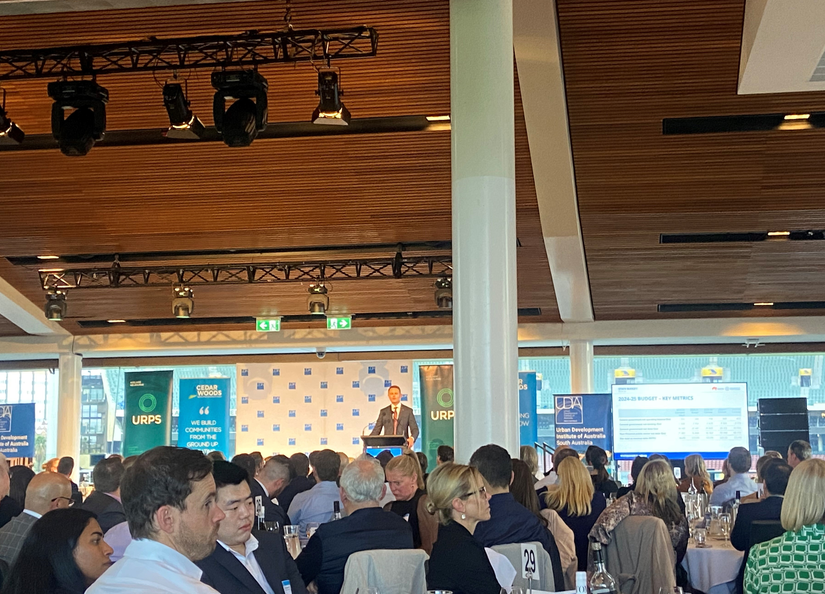 A large event filled with tables of people and the treasurer addressing the crowd