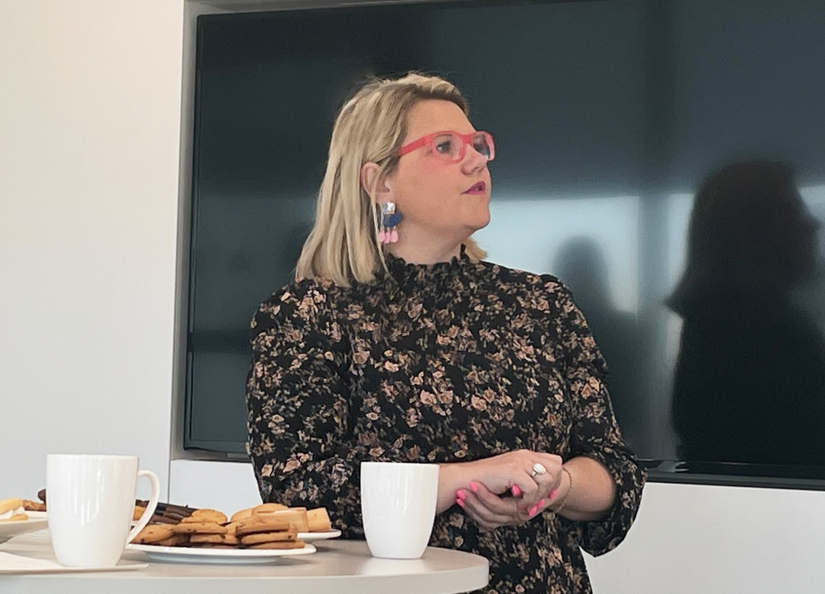 Woman in floral dress reading from a screen. In front of her is a table with coffee cups and biscuits.