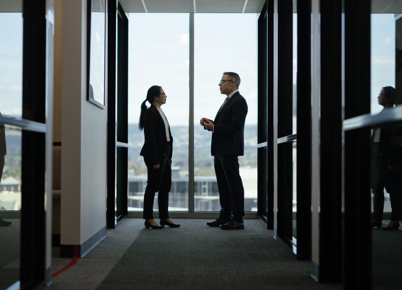 Young woman and older man dressed professionally having a conversation at the end of a corridor in front of a window with a view of the Adelaide Hills