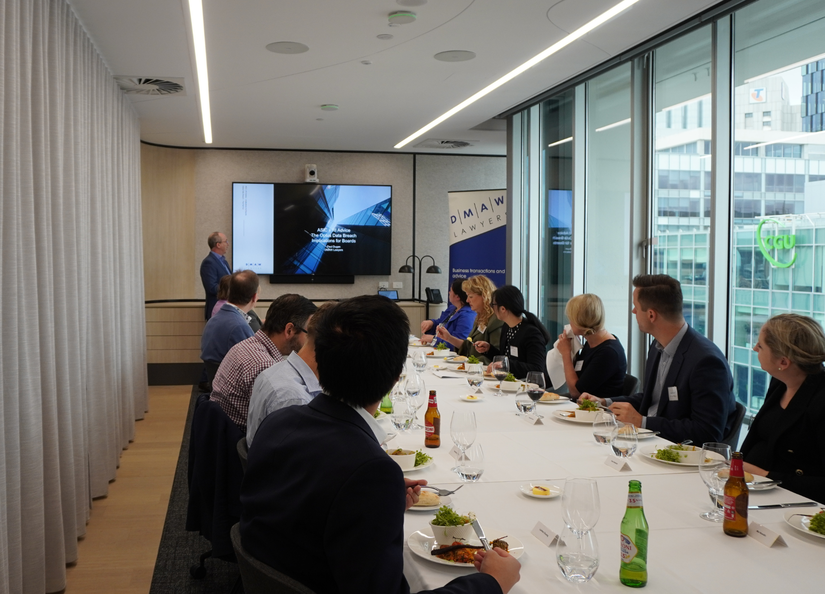 Group of men and women seated around a boardroom table eating lunch and watching a presentation