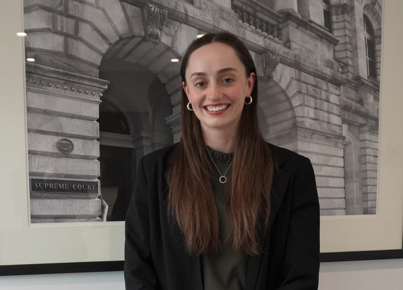 Young Caucasian female with long brown hair smiling for her photo, standing in front of a photo of law courts