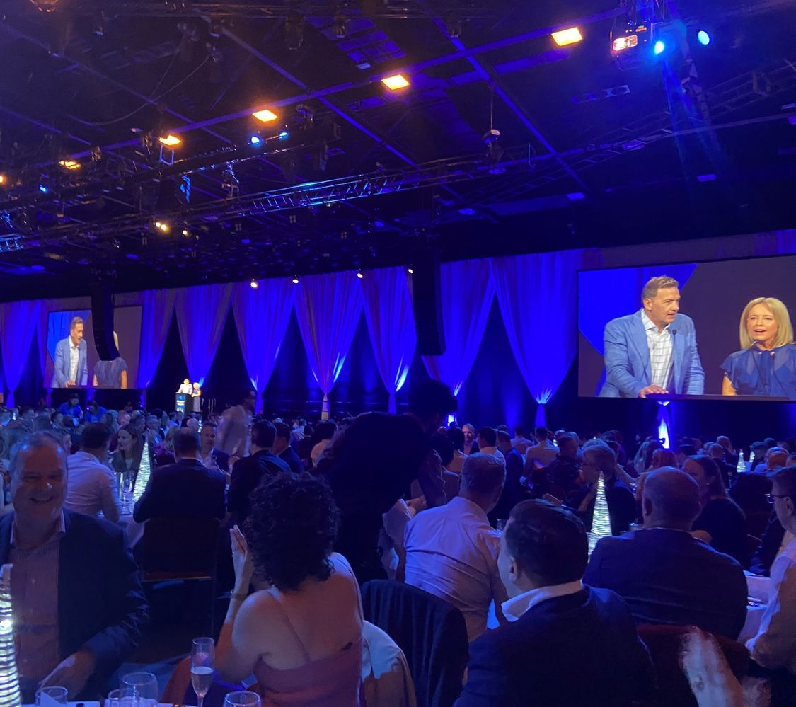 Large function room with people seated around round tables and presenters on big screens