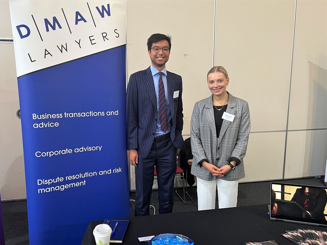 Young Asian man and Caucasian women in professional dress standing in front of a bright blue DMAW Lawyers Banner