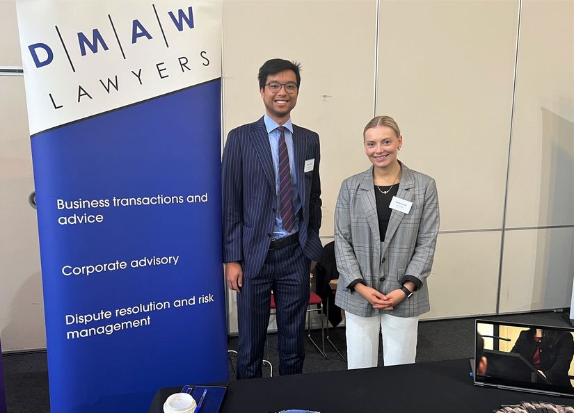 Young Asian man and Caucasian women in professional dress standing in front of a bright blue DMAW Lawyers Banner