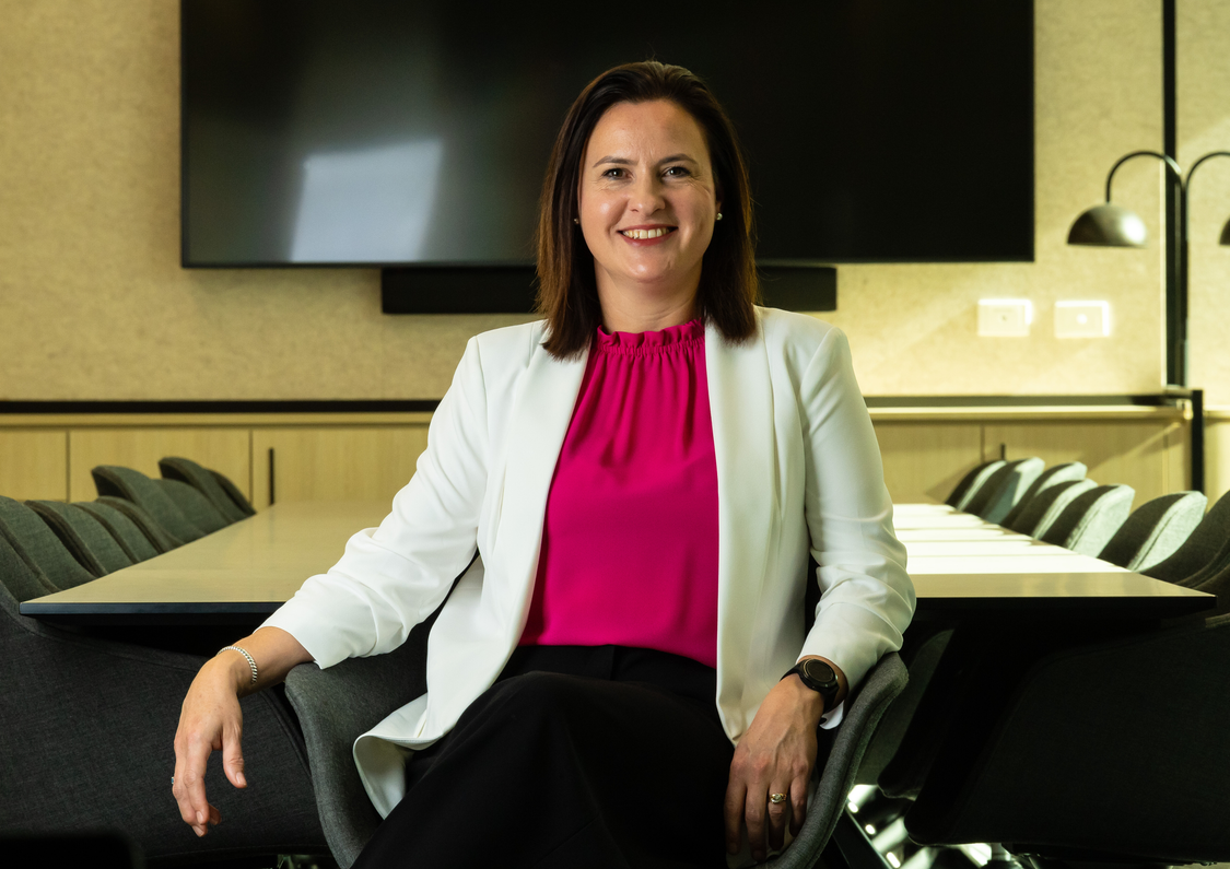 Chief Executive Officer Bernice Witkowski wearing a bright pink top and white blazer seated in a chair in the middle of a boardroom