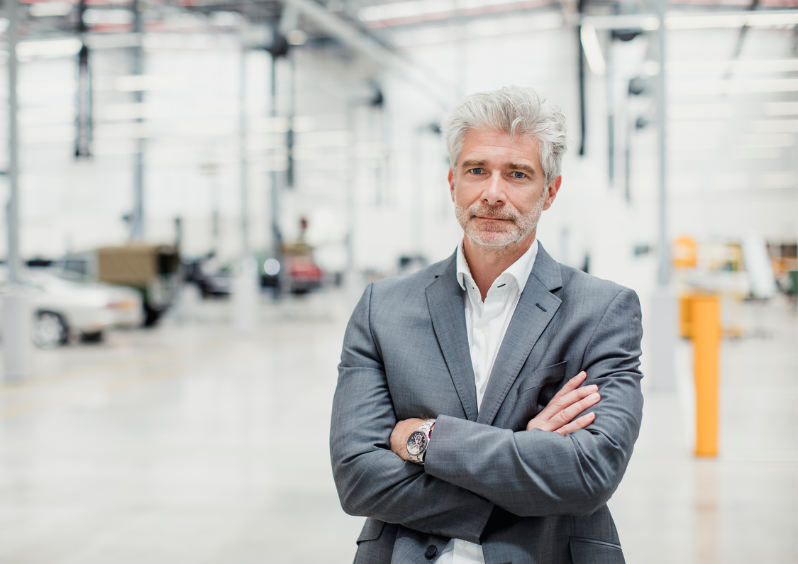 A middle aged man in a grey suit with arms folded, appears to be standing in a carpark