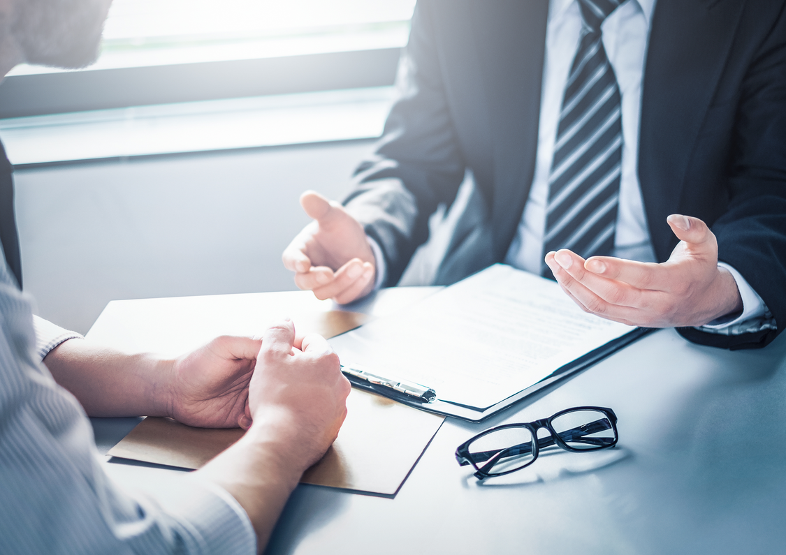 Two people discussing contracts. The contract is on the table with some reading glasses. The people are wearing business attire but only their hands can be seen