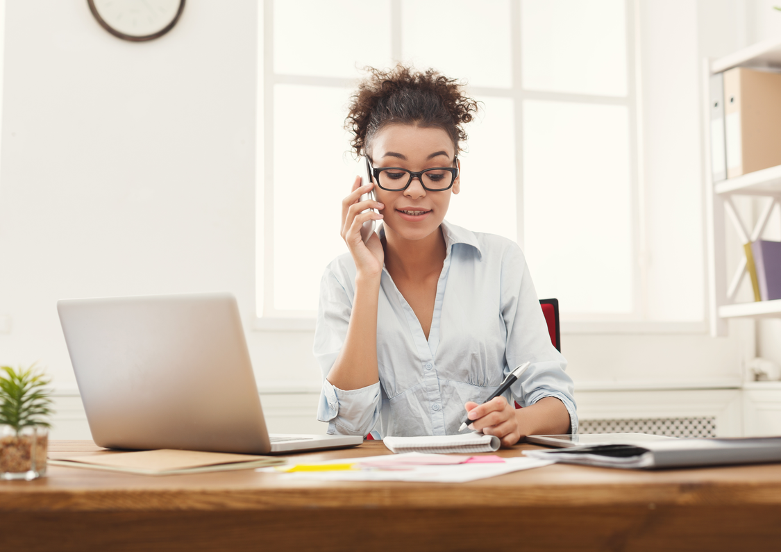 A young business woman on the phone at her desk. There is a laptop and other documents strewn across the desk.