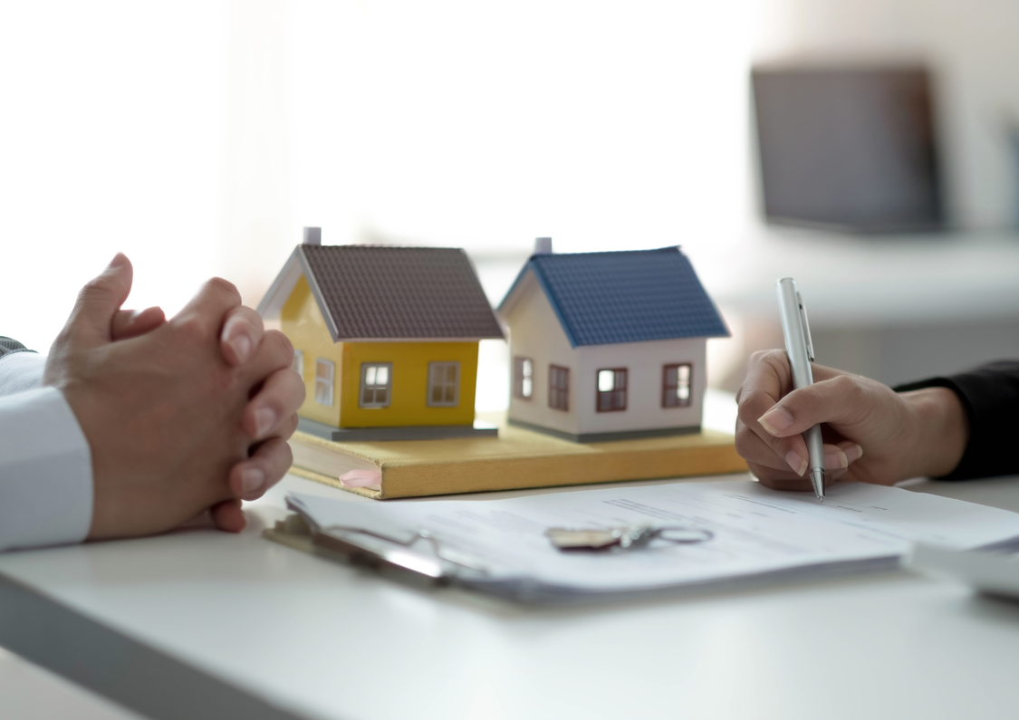 Two small houses on a table and hands on either side of the table. One set of hands holding a pen ready to sign a document.